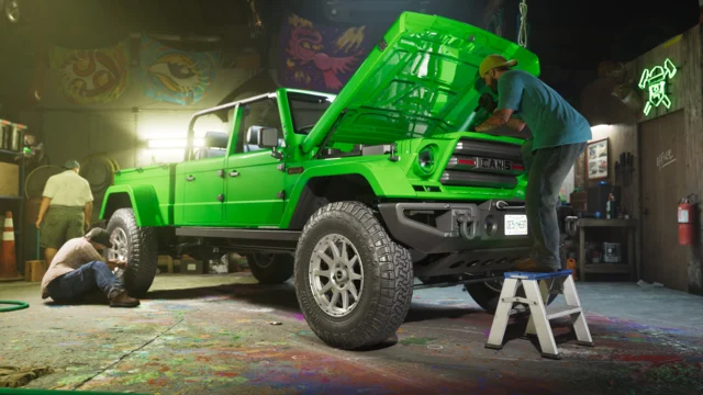 Inside an auto shop, two men are working on a green off-road vehicle with the hood up.